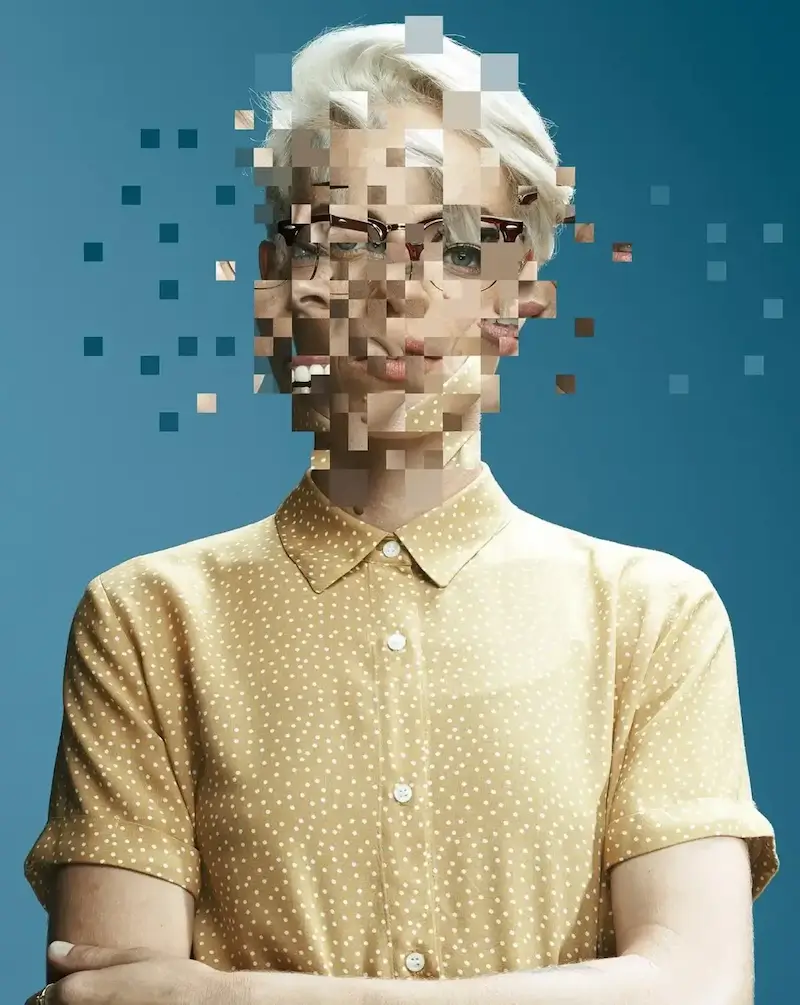 A person with cropped light hair and glasses stands with arms folded against a deep blue background, their face breaking apart into pixel squares.