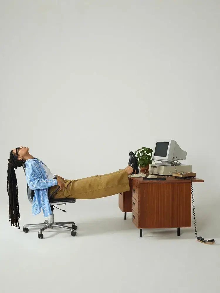 A person reclines right back in an office chair with their feet up on a desk holding a retro computer and a small plant.